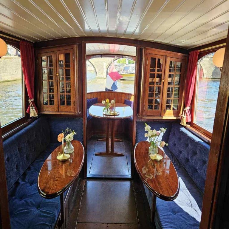 صالة طعام أنيقة داخل جولة عشاء بالقارب في فندق ريتز أمستردام مع طاولات خشبية وزهور طازجة وإطلالات على القناة