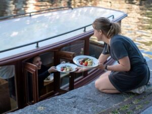 A server handing fresh plates of food from a canal-side stone quay directly to guests through the window of a classic Ams