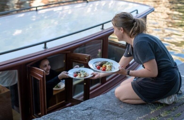 A server handing fresh plates of food from a canal-side stone quay directly to guests through the window of a classic Ams