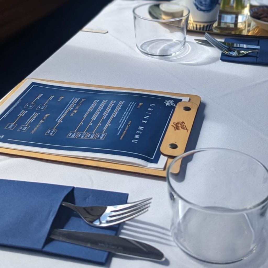 Elegant brunch table setting on an Amsterdam canal boat featuring a blue drink menu, silverware, and glassware.